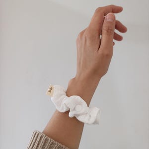 Puede incluir: Un coletero de tela blanca con un peque&ntilde;o cierre de bot&oacute;n a presi&oacute;n circular y una etiqueta rectangular de tela beige con el texto "Sir Co" impreso en negro. El coletero est&aacute; hecho de un material suave y fruncido para llevar en la mu&ntilde;eca.