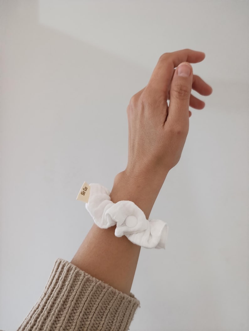 Puede incluir: Un coletero de tela blanca con un peque&ntilde;o cierre de bot&oacute;n a presi&oacute;n circular y una etiqueta rectangular de tela beige con el texto "Sir Co" impreso en negro. El coletero est&aacute; hecho de un material suave y fruncido para llevar en la mu&ntilde;eca.