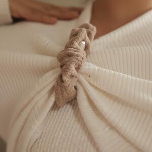 Puede incluir: Accesorio para el cabello de tela beige con un acento de perla, prendido a una parte superior crema acanalada. El accesorio est&aacute; torcido y anudado, creando una apariencia texturizada. La est&eacute;tica general es suave y elegante.