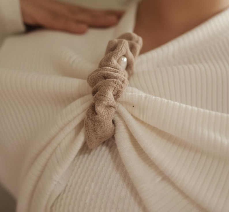 Puede incluir: Accesorio para el cabello de tela beige con un acento de perla, prendido a una parte superior crema acanalada. El accesorio est&aacute; torcido y anudado, creando una apariencia texturizada. La est&eacute;tica general es suave y elegante.