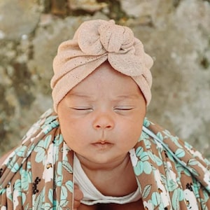 Könnte beinhalten: Ein Neugeborenes mit einem hellbraunen Turbanhut. Das Baby ist in eine braune Decke mit einem Blumenmuster in Blau- und Weißtönen gehüllt. Die Augen des Babys sind geschlossen.