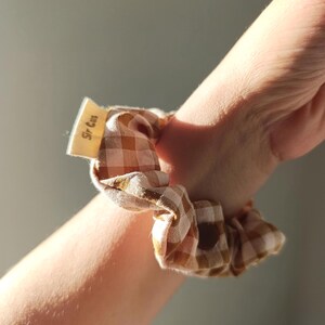 May include: A brown and white gingham scrunchie is wrapped around a wrist. The scrunchie has a small tag that reads "Sir Cus". The fabric is a light brown and white checkered pattern.