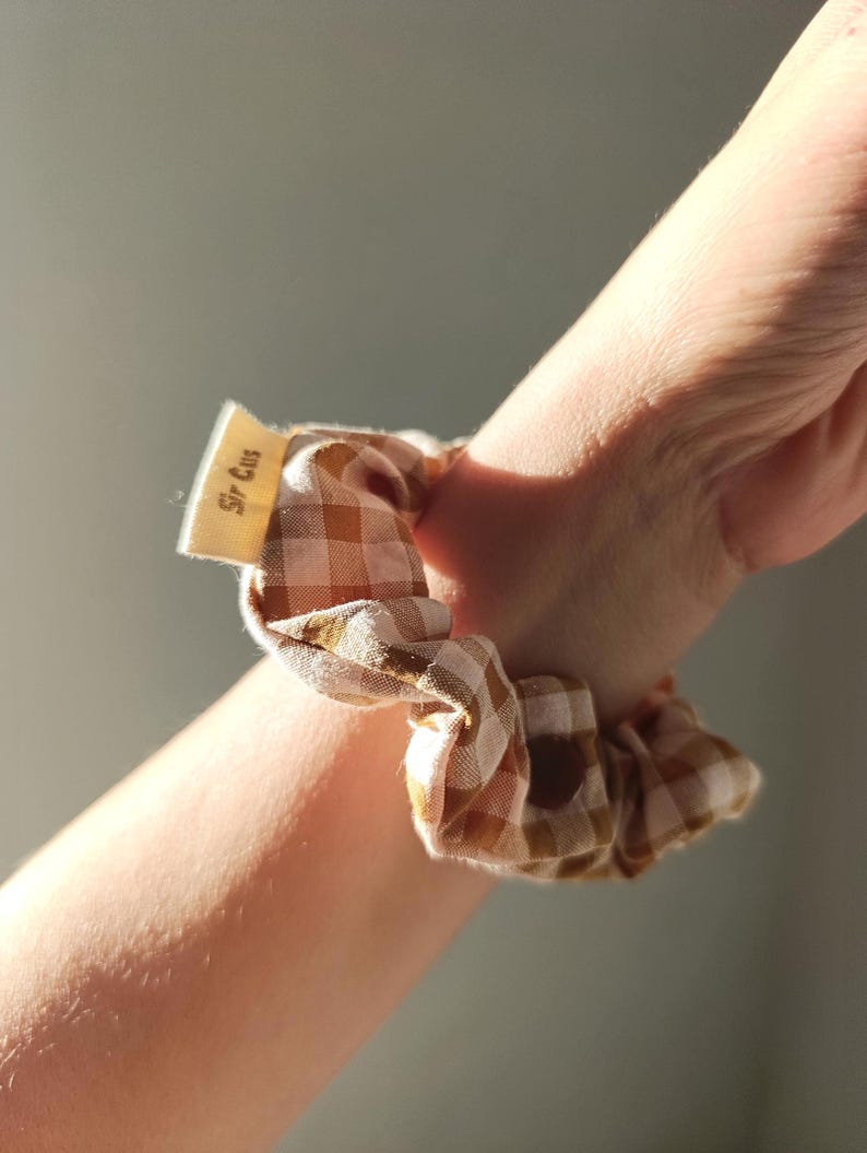May include: A brown and white gingham scrunchie is wrapped around a wrist. The scrunchie has a small tag that reads "Sir Cus". The fabric is a light brown and white checkered pattern.