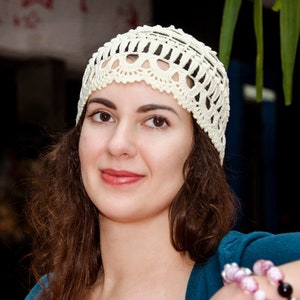 May include: A woman with long brown hair wearing a white crocheted hat. She is smiling and looking at the camera. She is wearing a blue shirt and has bracelets on her wrist.