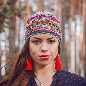May include: A woman wearing a colorful crocheted hat with red tassel earrings. She is wearing a black and white polka dot shirt.