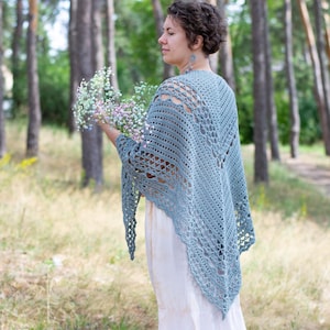 Könnte beinhalten: Eine Frau trägt einen hellblauen Häkel-Schal mit einem weißen Kleid und braunen Schuhen. Sie steht in einem Wald und hält einen Strauß kleiner weißer Blumen.