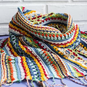 A scarf in primary colors with a stunning design. It is handmade and placed against the backdrop of a white brick wall.