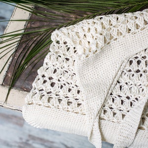 Könnte beinhalten: Ein weißer Häkelcardigan mit einem zarten Lochmuster. Der Cardigan ist gefaltet und auf einer Holzoberfläche mit einem grünen Palmblatt gelegt.