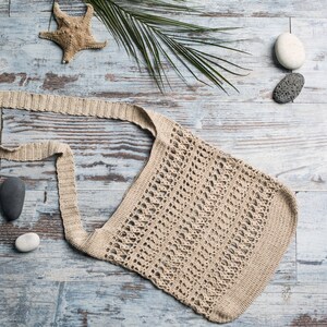 May include: A beige crocheted tote bag with a long strap, laying on a light blue wooden surface with a starfish, palm leaves, and stones.