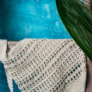May include: A close-up of a beige crocheted tote bag with a diamond pattern. The bag is hanging on a wooden shelf against a blue background.