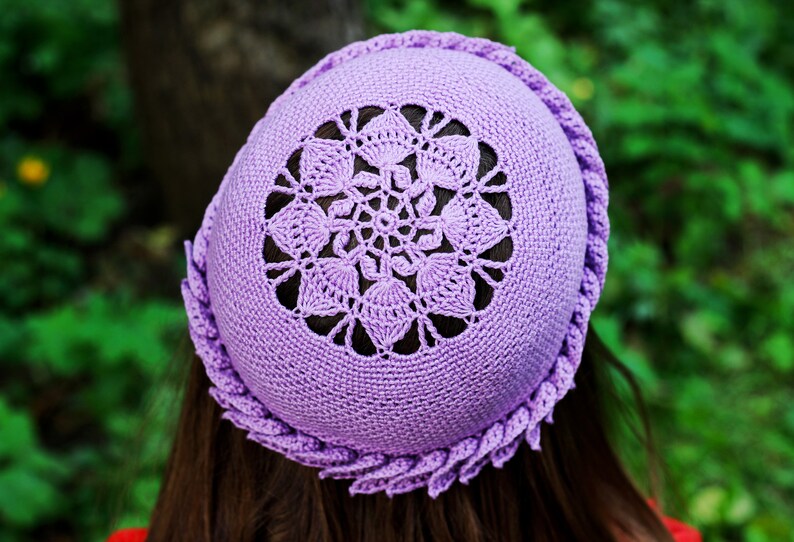 A purple crocheted hat with a delicate floral design on a top side of the beanie. The hat is made with a soft, lightweight yarn and has a comfortable fit.