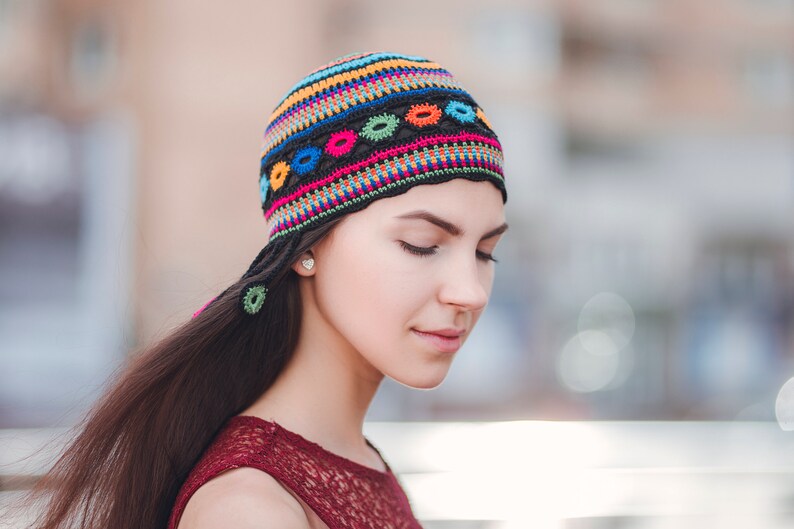 May include: A woman with long dark hair wearing a colorful crocheted hat with a black background and colorful geometric designs. The hat has a long tail that hangs down her back.