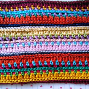 K&ouml;nnte beinhalten: Mehrfarbiger H&auml;kelstoff mit Streifen in verschiedenen Farben und Texturen. Der Stoff ist aus verschiedenen Garnfarben hergestellt, darunter Rot, Blau, Gr&uuml;n, Gelb und Lila.