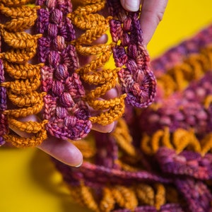 May include: A close-up of a crocheted scarf in shades of orange and purple. The scarf is made with a delicate stitch pattern and has a soft, textured feel.