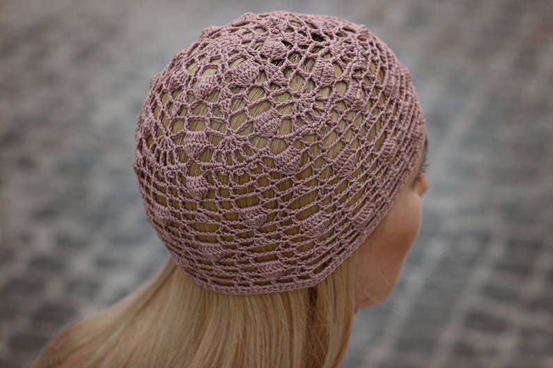 May include: A close-up of a crocheted hat in a dusty rose colour. The hat is made with a delicate openwork pattern.