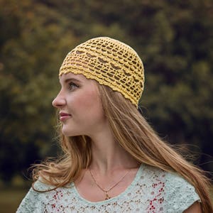 Profile of a woman's head against a background of green bushes. The woman is wearing a crocheted openwork lace hat made from fine yarn.