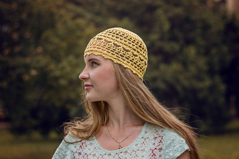 Profile of a woman's head against a background of green bushes. The woman is wearing a crocheted openwork lace hat made from fine yarn.