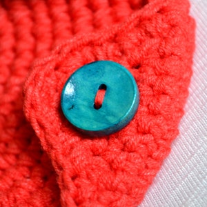 May include: A close-up of a teal blue wooden button sewn onto a red crocheted fabric.