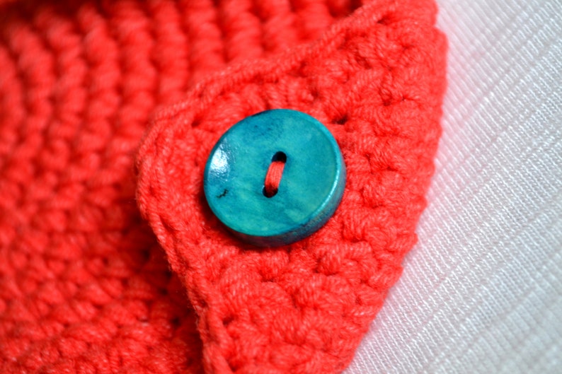 May include: A close-up of a teal blue wooden button sewn onto a red crocheted fabric.