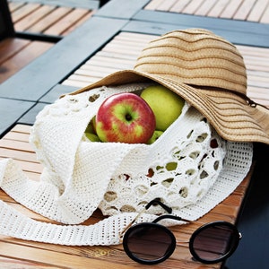 May include: A white crocheted tote bag with a brown straw hat on top. The bag is filled with green and red apples. A pair of black sunglasses are lying on the wooden table next to the bag.