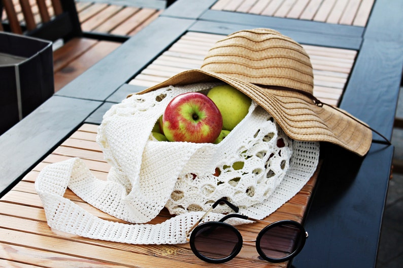 May include: A white crocheted tote bag with a brown straw hat on top. The bag is filled with green and red apples. A pair of black sunglasses are lying on the wooden table next to the bag.