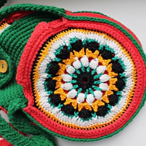May include: A crocheted bag with a circular design in red, green, white, black, and yellow. The bag has a long strap and a button closure.