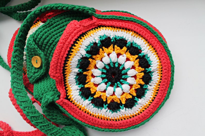 May include: A crocheted bag with a circular design in red, green, white, black, and yellow. The bag has a long strap and a button closure.