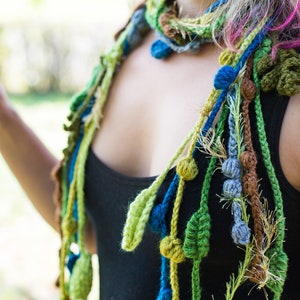May include: A close-up of a woman wearing a colorful crocheted scarf with green, blue, and brown accents. The scarf features crocheted leaves and beads.
