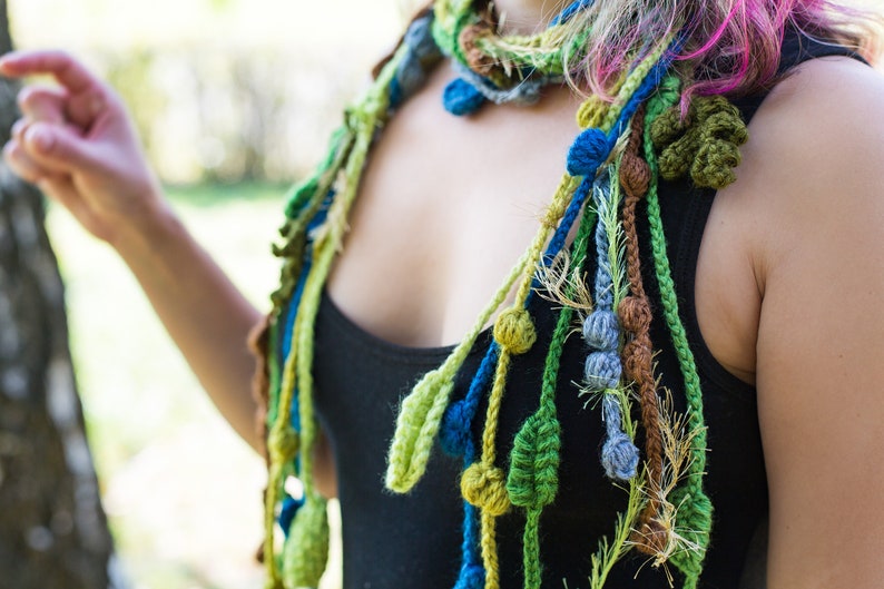 May include: A close-up of a woman wearing a colorful crocheted scarf with green, blue, and brown accents. The scarf features crocheted leaves and beads.