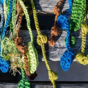 May include: A collection of crocheted strands in various colors, including green, blue, brown, and white, with small crocheted balls attached. The strands are hanging from a wooden surface.