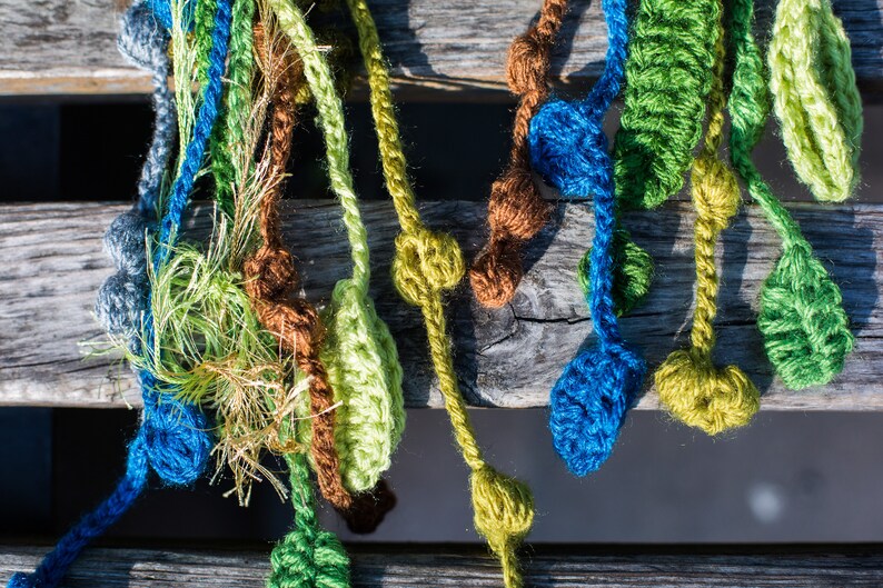 May include: A collection of crocheted strands in various colors, including green, blue, brown, and white, with small crocheted balls attached. The strands are hanging from a wooden surface.