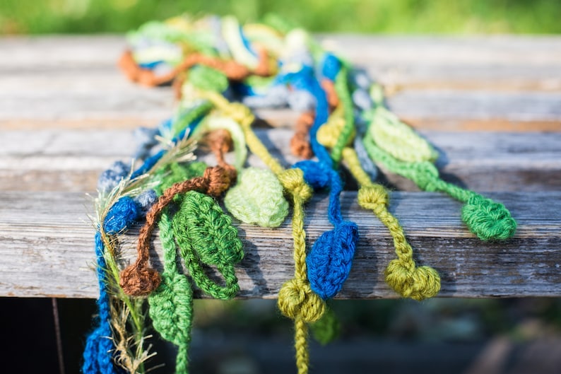 May include: A crocheted scarf with green, blue, brown, and yellow leaves. The scarf is made of yarn and is a handmade craft.