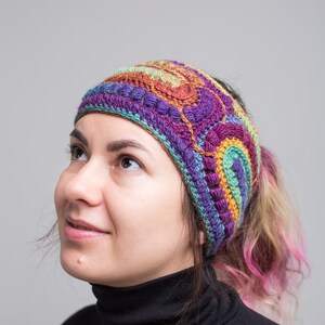 May include: A woman wearing a colorful crocheted headband with a multi-colored design. The headband is made of yarn in shades of purple, green, orange, and brown. The woman is wearing a black turtleneck sweater.