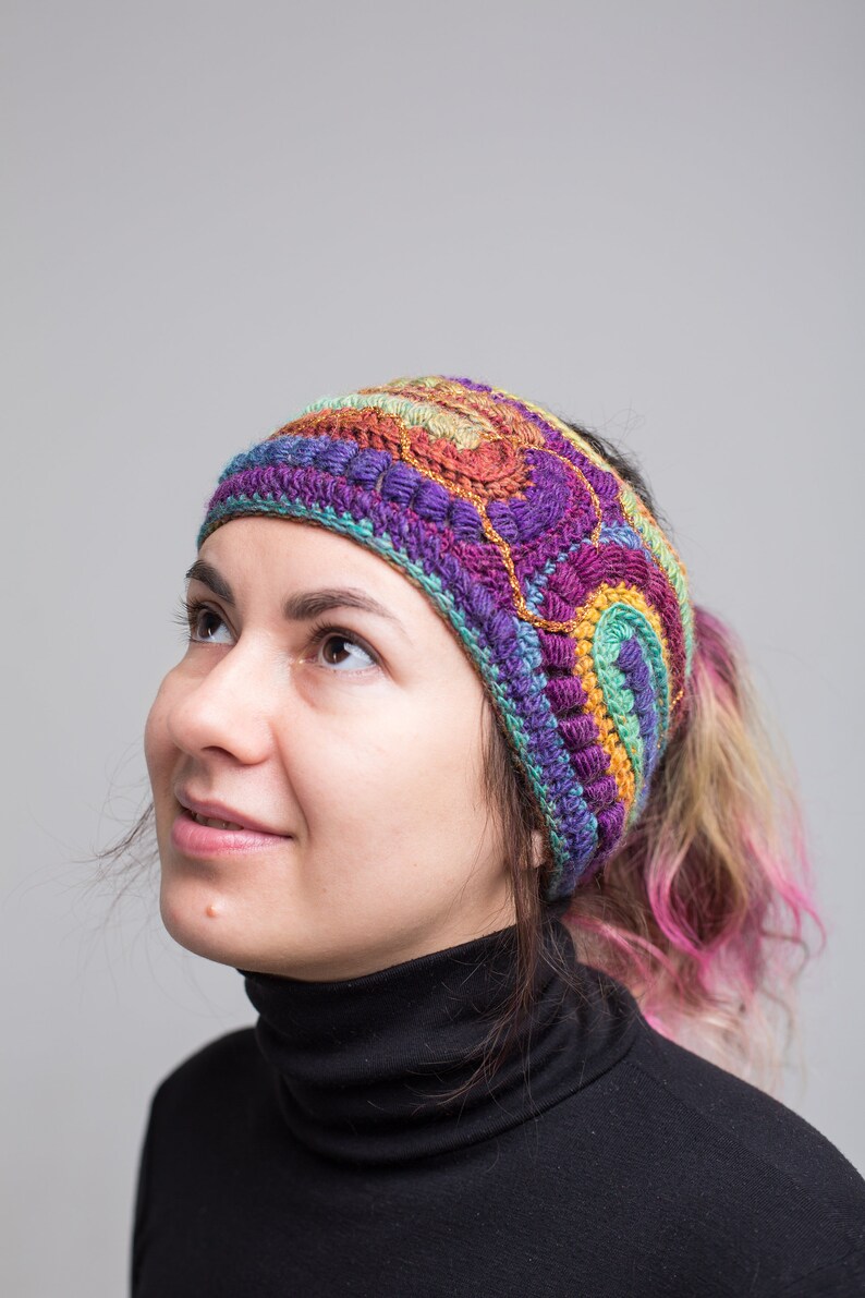 May include: A woman wearing a colorful crocheted headband with a multi-colored design. The headband is made of yarn in shades of purple, green, orange, and brown. The woman is wearing a black turtleneck sweater.