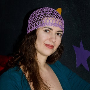 May include: A woman with long brown hair wearing a purple crocheted hat and a teal blue cardigan.