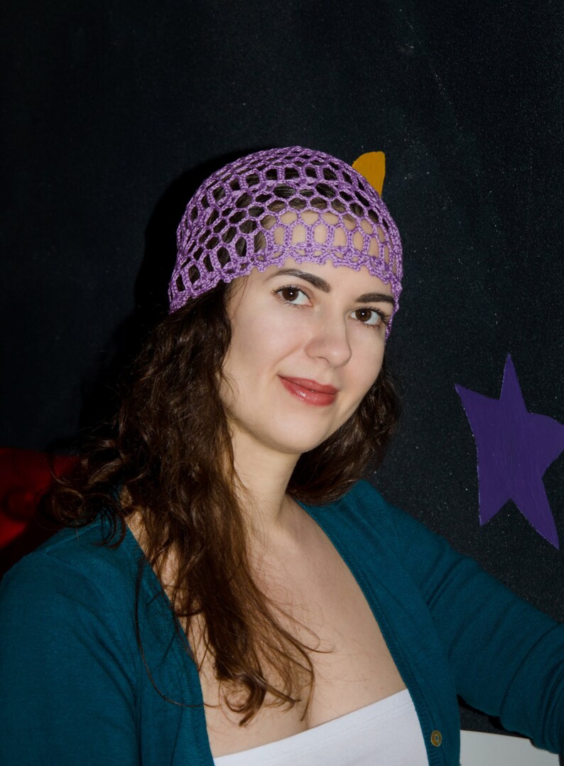 May include: A woman with long brown hair wearing a purple crocheted hat and a teal blue cardigan.