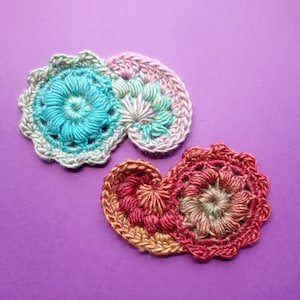 May include: Two crocheted flower motifs in a heart shape. One motif is pink, green, and blue, and the other is red, orange, and brown.