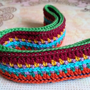 K&ouml;nnte beinhalten: Bunt geh&auml;keltes Band mit gr&uuml;nem Rand. Das Band ist aus verschiedenenfarbigen Garnen gefertigt, darunter Rot, Orange, Gelb, Blau und Lila.