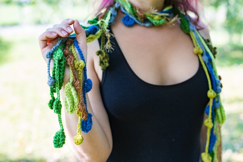 May include: A crocheted necklace with green, blue, and brown yarn. The necklace has a long, flowing design with multiple strands and beads.