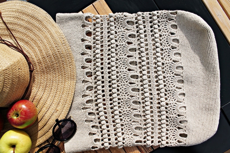 May include: A beige crocheted tote bag with a woven pattern. The bag is lying on a wooden surface next to a straw hat and apples.