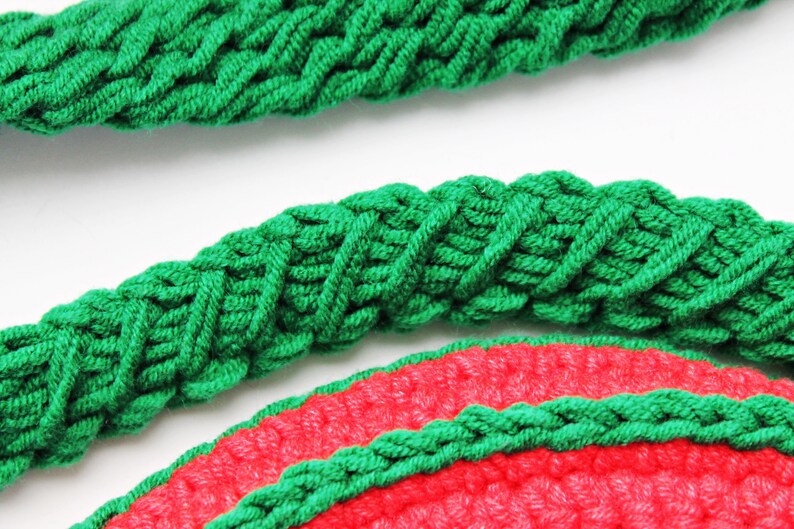 May include: Close-up of a green crocheted rope with a braided pattern. The rope is wrapped around a red crocheted circle.
