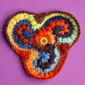 Op de afbeelding: Een gehaakt hartvormig triquetra-symbool met een veelkleurig ontwerp in tinten oranje, bruin, blauw, groen en geel.