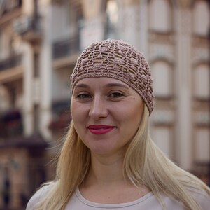 May include: A woman with blonde hair is wearing a light brown crocheted hat. She is smiling and looking at the camera.