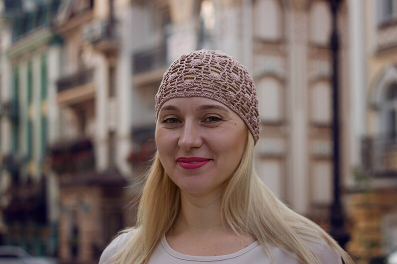 May include: A woman with blonde hair is wearing a light brown crocheted hat. She is smiling and looking at the camera.