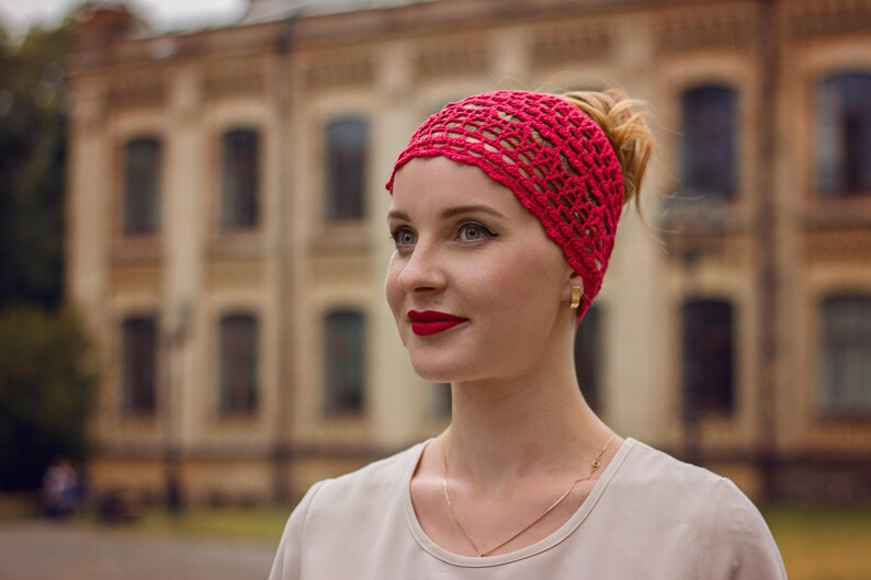 May include: A woman with a red crocheted headband and a white shirt. She is looking to the side and smiling.