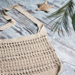 May include: A beige crocheted tote bag with a long strap, lying on a light wood surface with a starfish, palm leaves, and stones.
