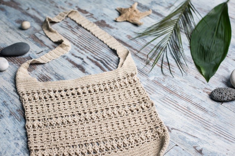 May include: A beige crocheted tote bag with a long strap, lying on a light wood surface with a starfish, palm leaves, and stones.