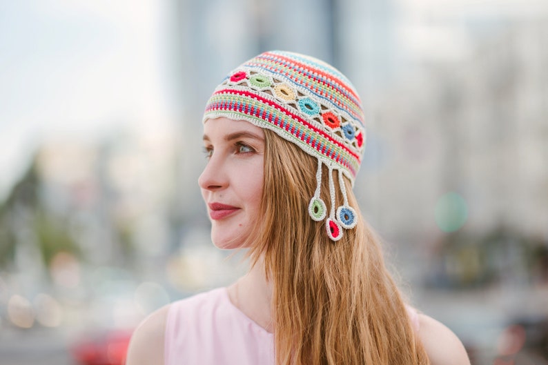 May include: A woman wearing a white crocheted hat with colorful stripes and tassels. The hat has a geometric pattern with red, blue, green, and yellow accents.
