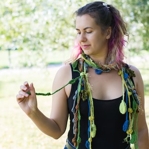 May include: A woman wearing a black tank top and a long, colorful crocheted scarf with green, blue, and yellow accents. The scarf is made of yarn and has a variety of textures and shapes.