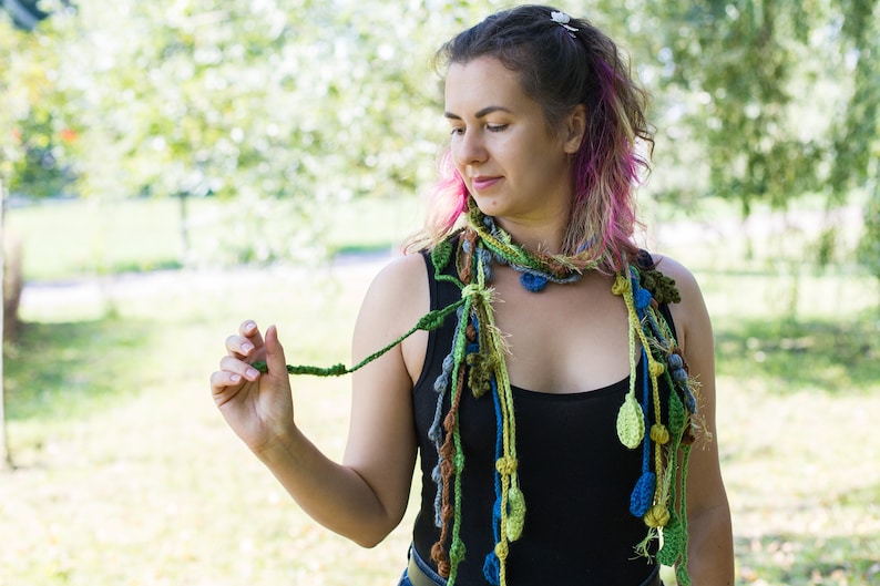 May include: A woman wearing a black tank top and a long, colorful crocheted scarf with green, blue, and yellow accents. The scarf is made of yarn and has a variety of textures and shapes.